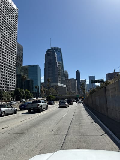 This media file is a vertical photograph that captures a dynamic urban scene from a freeway in Los Angeles, California, under clear, bright daylight. When viewed in its intended landscape orientation (rotated 90 degrees counter-clockwise), it shows a multi-lane concrete freeway winding through a bustling city, flanked by towering skyscrapers and urban greenery.
The freeway in the midground and foreground is populated with numerous cars, suggesting moderate to heavy traffic. Vehicles visible include a white pickup truck, a dark sedan, a silver Mercedes-Benz sedan, and a white MINI Cooper convertible, among others. The road features clear white lane markings and reflective studs.
On the left side of the freeway, a dense cluster of modern high-rise buildings forms the impressive city skyline. Notable structures include a prominent skyscraper with a spire, featuring the logos "pwc" and "DMC." Another large, reflective glass building clearly displays "SMBC MANUBANK" on its facade. To the left, a building with a distinctive white grid pattern is visible, and another structure bears the partially legible text "CATERRESS." Interspersed between the buildings and alongside the freeway are palm trees and other green foliage.
The sky is a vivid, cloudless blue, indicating excellent weather conditions. Strong sunlight casts sharp shadows, confirming it is daytime, likely morning or early afternoon. Key location cues are provided by green freeway signs: one reads "110 Harbor Fwy," and another partially visible sign indicates "7th & 8th Sts Exit," firmly placing the scene in downtown Los Angeles. A license plate "9DG6549" is visible on the white pickup truck.