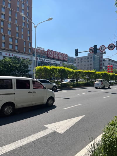 This image depicts a street scene in Changchun City, China, likely on a sunny day given the bright lighting and clear blue sky. The foreground is dominated by a white van driving on the right side of a multi-lane asphalt road. A large white arrow indicating a forward direction is painted on the road surface. To the left of the van, another white car is partially visible, also driving in the same direction.
In the background, a modern, multi-story building with a brick facade and numerous windows stands prominently on the left side of the street. Next to it is a building with signage in Chinese, including "BANK OF CHINA." Across the street, a line of lush green trees forms a median or buffer zone. Beyond the trees, more buildings are visible, including a taller structure with glass facades and architectural details.
Traffic signals are present, with one showing a green light for straight movement and a red light for turning. Traffic signs indicating a speed limit of 50, a left turn restriction, and a motorcycle prohibition are also visible. The overall environment suggests a typical urban setting with commercial buildings and well-maintained infrastructure. There are no people clearly visible in the image.