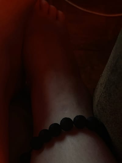 This is a close-up, low-light photograph focusing on a person's foot and ankle, adorned with a black beaded bracelet. The subject's foot, with visible toes, is positioned on the left side of the frame, bathed in a warm, orange-toned light. To the right, the ankle is wrapped with a dark, textured bracelet composed of multiple spherical beads. A shadowed, textured surface, possibly fabric or a rough ground, lies behind the foot and ankle, creating a sense of depth. The overall lighting is dim, with strong shadows obscuring parts of the scene, suggesting an indoor setting at night or in a dimly lit environment. No text is visible, and no specific activity or event is clearly depicted, other than the presence of the individual. The focus is on the texture and form of the foot, ankle, and bracelet, with the warm light creating a potentially intimate or relaxing mood. The location context of Podolsk, Russia, is noted, but no specific visual cues within the image confirm this.