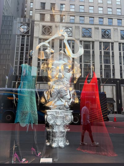 This media file captures a street view through a window display, likely in a bustling urban environment. The foreground is dominated by a striking, large-scale silver sculpture that appears to be a stylized human face, possibly with classical architectural elements integrated into its design. The sculpture is partially obscured by reflections on the glass, creating a layered effect.
Behind the sculpture, a vibrant red, string-like art installation is prominently displayed. A person, likely a man, is visible within this red installation, wearing a light-colored t-shirt, dark shorts, and sneakers. He is partially silhouetted and appears to be walking or posing within the artwork. The red installation is semi-transparent, allowing glimpses of the background.
The background reveals a tall building with numerous windows, reflecting the sky and surrounding structures. Further back, a dark blue bus is visible, with some text on its side, including what appears to be "BROADWAY" in reverse. Another smaller structure with red and yellow diagonal stripes is also present.
The lighting suggests it could be late afternoon or early evening, with the sun casting long shadows and creating a golden hue on some surfaces, as seen on the silver sculpture. The scene evokes a sense of vibrant city life and artistic expression. The presence of a bus and the architectural style of the building suggest a location in a major city, consistent with the provided location context of New York, USA, specifically Broadway.