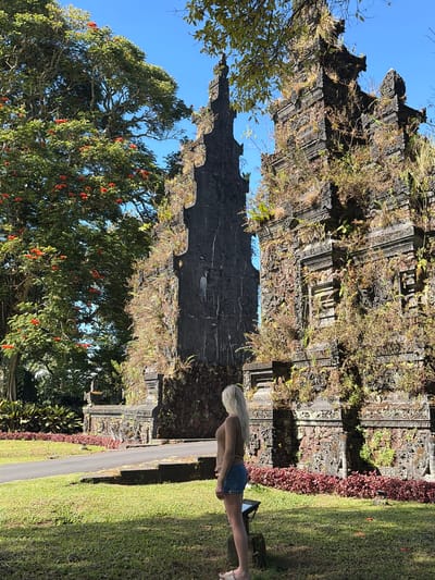 A young woman with blonde hair, wearing a brown long-sleeved shirt and denim shorts, stands facing a large, ancient, moss-covered stone archway in the city of Sukasada, Indonesia. The archway is ornately carved and covered in lush green vines and plants. To the left of the archway, a large tree with vibrant red flowers is in bloom. The ground is covered in green grass, and a paved pathway leads up to the archway. The sky is a clear, bright blue.