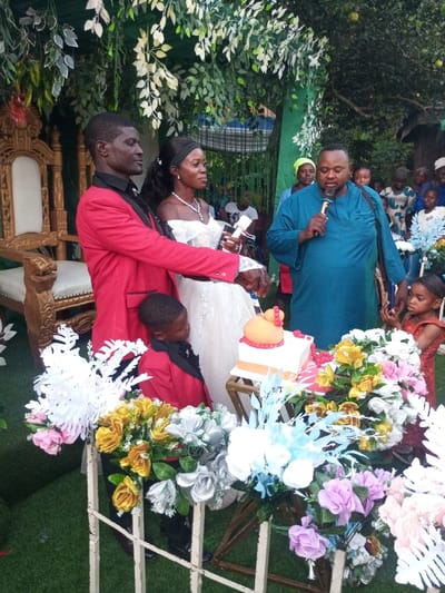 The image captures an outdoor celebratory event in Amokvo, Nigeria, set on artificial green turf with a backdrop of green and white artificial foliage and natural trees. At the center, a dark-skinned adult male in a red blazer and a dark-skinned adult female in a white off-the-shoulder wedding gown are jointly cutting a two-tiered cake with a white-handled knife. The cake has a white rectangular base with gold stripes and an orange-red round top tier adorned with red beads. A young dark-skinned boy in a red blazer stands near the groom, observing the cake. To the right, a dark-skinned adult male in a teal traditional garment holds a gold microphone, addressing the scene. An ornate gold throne chair with white upholstery is visible on the far left. Several adult and child guests observe from the background. The foreground features numerous artificial flower arrangements in white, gold, silver, pink, blue, and purple.