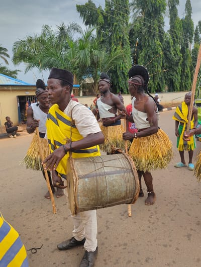 Several people are on a street in what appears to be a parade or celebration in Maigizo, Nigeria. At the forefront, a man wearing a black cap, a white shirt, a yellow and black striped garment, and off-white pants is holding a drum that he is playing. Another man behind him wears a white garment and a straw skirt. He is holding a wooden instrument and is barefoot. A third man with similar attire follows behind the second man. The setting is a paved street with trees in the background. There are buildings in the background, and the overall environment indicates a city street.