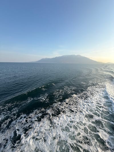 The scene depicts a marine environment near Palaiopoli, Greece. In the foreground and mid-right, a prominent white wake of churned water extends, indicating the passage of a vessel. The majority of the midground consists of dark blue-green sea water with visible surface ripples. Across the horizon, a large landmass featuring mountains with subtle lighter-colored peaks is present. The sky above is clear blue, with a faint yellowish glow on the far right, suggesting either sunrise or sunset. No people or specific objects beyond the natural environment and vessel wake are visible.