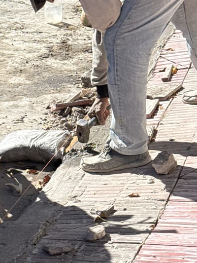 A person wearing light-wash jeans and grey canvas shoes is standing outdoors on a cracked, light-colored concrete surface. The person is using a hammer to break a stone. A small, clear plastic container sits to the left of the hammer. The location appears to be near a building with a textured wall, partially obscured by shadow, featuring vertical stripes that alternate between light grey and reddish-brown. The photo was taken in Salta, Argentina.