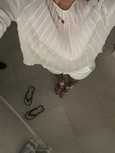 The image shows the feet and legs of a person wearing white sandals, white shorts, and a white, striped shirt. The person is standing in a tiled bathroom, with two pairs of black flip-flops lying on the floor. The bathroom is located in Miami Beach, Florida, United States, at the coordinates 25.773 latitude and -80.14 longitude. The scene is a simple bathroom, and the person appears to be preparing to leave.