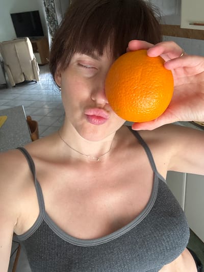 A rotated, medium shot depicts a woman in a gray tank top holding an orange up to her face. She is indoors, in a kitchen setting. Her eyes are closed and she is puckering her lips towards the orange. She has short, dark brown hair with bangs. A gold necklace is visible around her neck. Her left hand is holding the orange. The background includes a gray countertop, tiled wall, a brown paper bag, and a black cabinet. The location is not discernible from the provided image.