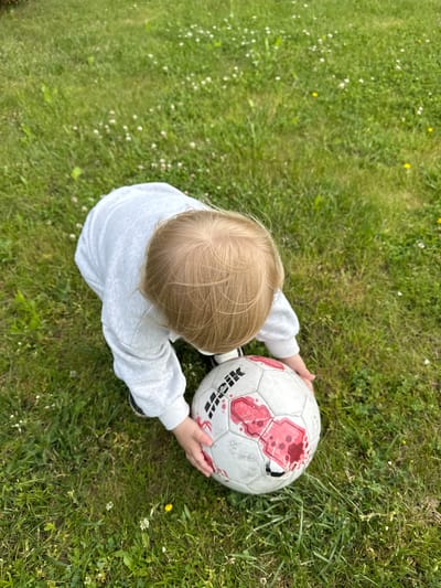An overhead shot shows a young child with blonde hair, wearing a light gray sweatshirt, kneeling on a green grassy lawn dotted with small white clover flowers. The child is reaching for a white soccer ball with red markings. The ball appears to be slightly worn and has a small rip on one of its panels. The scene suggests an outdoor activity in a park or backyard. There are no distinguishing landmarks in the image to confirm its specific location within Gvardeysk, Russia.