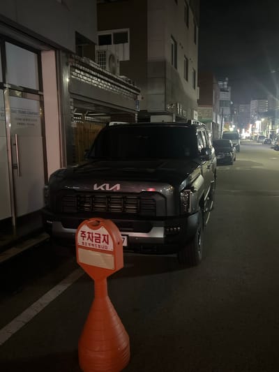 The image shows a dark-colored Kia SUV parked on a city street at night, in Gwangju, South Korea. The vehicle is positioned in the foreground, facing the viewer, with its front grille and headlights clearly visible. A red and orange traffic cone with Korean text, "주차금지" (No Parking), is placed in front of the SUV. The street is paved and appears to be a typical urban setting with buildings lining one side of the road. Other vehicles are visible further down the street, and streetlights illuminate the scene. The lighting suggests it is nighttime. No people are visible in the image.
