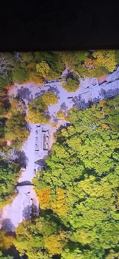 The image is an aerial view of a park-like area with a path winding through trees. On the path, there appears to be a ruin or structure made of stone blocks, possibly the remains of an old building. The trees display autumn foliage, with a mix of green, yellow, and orange leaves. The scene is likely captured during the daytime. Based on the visual cues, this location could be the Dendropark "Alexandria" in Bila Tserkva, Ukraine, known for its scenic landscapes, ancient trees, and historical structures.