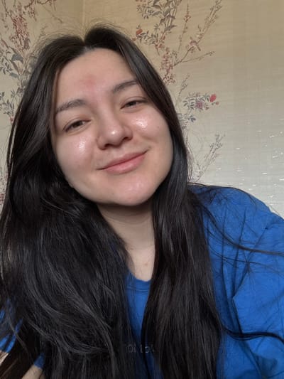 A female individual is depicted in a chest-up portrait, positioned indoors in the city of Kurovskoye, Russia. She has long, dark, straight hair, parted centrally, framing her face and falling over her shoulders. Her face is oriented slightly to her left, looking towards the camera with a gentle, closed-mouth smile. Her skin appears to have a natural sheen, and a singular reddish mark is present on her forehead above the nasal bridge. She is wearing a blue, loose-fitting garment, possibly a t-shirt, with a faint, partially obscured light-colored inscription visible on its front. The background consists of a light-colored wall covered with wallpaper featuring a repeating pattern of dark floral branches, red flowers, green leaves, and small dark birds.