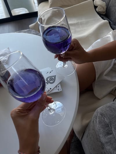 In Baan Tai, Thailand, two glasses of purple liquid are held by unseen hands over a white table. A deck of playing cards, including two aces of spades and a two of spades, lies on the table. The scene appears to be indoors, possibly in a living room, with soft seating and a reflective surface visible in the background.