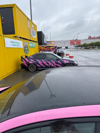 A purple sedan with a pink lightning bolt decal is parked outdoors in Shushary, Russia, under an overcast sky. The car is parked next to a bright yellow shipping container with a black door and a red rope attached. Another black shipping container is visible behind the yellow one. In the background, a large building with red and white signage, including "SUNLIGHT" and what appears to be "Гипермаркет" (Hypermarket), stands across a wet asphalt parking lot. Several other cars are parked in the lot. The foreground shows the roof of another vehicle, which is dark and wet, with a pink stripe visible along its edge. The ground is covered in puddles, indicating recent rain. There are no people clearly visible in the frame.