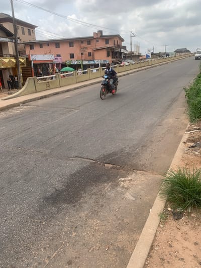 A man rides a motorcycle on a paved road in Shagamu, Nigeria. The road has several cracks and is lined with a concrete barrier. Buildings with shops are visible on the left side of the road, and lush green vegetation grows on the right. The sky is cloudy.