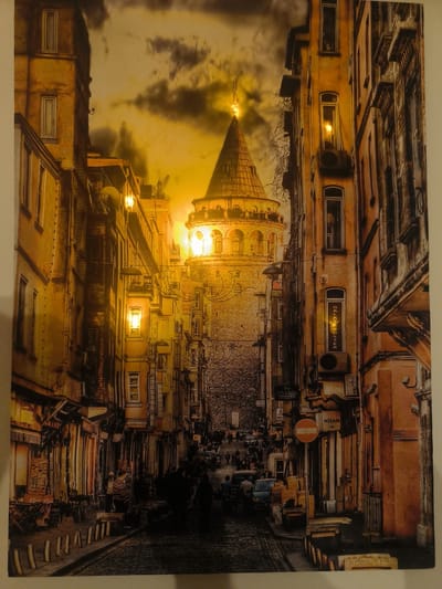 The image depicts a street scene with the Galata Tower in the background, suggesting a location in Istanbul, Turkey, not Coirac, France. The tower, a prominent landmark, is centrally located and illuminated, with a conical roof and ornate detailing. The foreground is dominated by a narrow cobblestone street lined with old buildings on either side. These buildings are multi-story, with many windows and architectural details such as balconies and awnings. The facades are predominantly yellow and orange, giving the scene a warm, aged feel.

The street is populated by a number of people, mostly silhouetted and indistinct, suggesting a bustling urban environment. Some are walking, while others appear to be standing or engaged in activities along the street. There are also vehicles, including a blue car and a van, and various objects such as street lamps, signs, and what appear to be market stalls or storefronts. The lighting suggests it is either dusk or dawn, with warm tones from the setting or rising sun filtering through the cloudy sky. The overall impression is one of a vibrant, historic city street.
