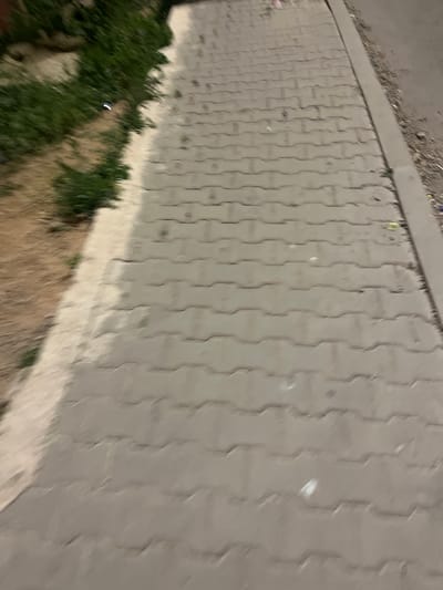 A ground-level, oblique angle image depicts a paved area consisting of interlocking concrete blocks. A strip of green vegetation borders the paved area on the left, transitioning into a patch of dry, brown earth. A concrete curb runs along the top of the paved area. The image appears to be taken at night or in low light conditions. The exact location within the city of Ouaouizert, Morocco, cannot be determined without further information.