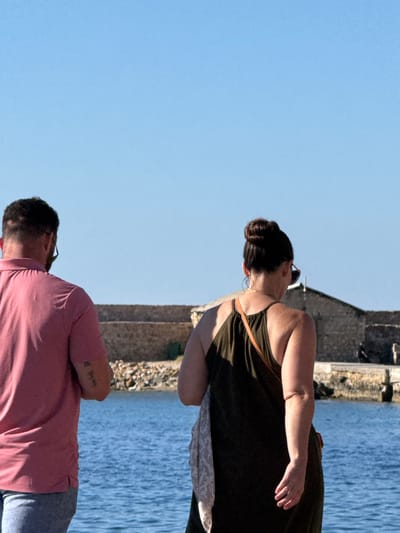 The image shows two individuals standing on a paved area, facing away from the camera, with a body of water in the foreground and a stone building with a blue sky in the background. The person on the left is wearing a pink polo shirt and jeans. The person on the right is wearing a dark olive green dress and a light-colored scarf draped over their arm. Both individuals are wearing sunglasses. The stone building appears to be historical, with weathered stones and a textured facade. The water is a vibrant blue, reflecting the clear sky. The scene is set in Chania, Greece, likely near the Venetian harbor area, given the architectural style of the building and the presence of the sea.