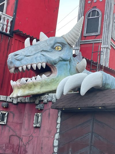 This media file features a close-up, low-angle shot of a large, decorative dragon head sculpture emerging from a red building. The dragon, with its light blue and green scales, prominent white teeth, and yellow eye, appears to be bursting through the structure. Several large, white tusks or horns are visible above its head, adding to its fearsome appearance.
The building itself is painted a vibrant red and appears to be made of panels that are artificially cracked to suggest an aged or damaged facade. There are decorative elements on the building, including what looks like a small, arched window with grey trim and a barred opening. Along the side of the building, there are sections of simulated stone blocks. Small, yellow lights are embedded in the wall near the stone blocks, suggesting these might be decorative lighting fixtures or part of the theme. A dark, textured roof-like structure extends from the top of the building, with simulated shingles.
The overall impression is of a theatrical or themed attraction, possibly a facade for a shop, restaurant, or amusement park. The image is brightly lit, suggesting it was taken during the daytime. There is no visible text in the image. The scene evokes a sense of fantasy and perhaps a touch of playful scariness. The orientation of the photo is such that the dragon's head is at the bottom, and the building extends upwards.
