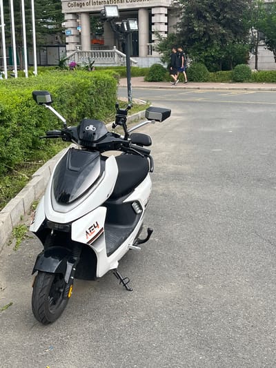 The image depicts an outdoor scene at the College of International Education in Changchun City, China. In the left foreground, a white and black electric scooter, bearing "olio" and "AEY" branding, is parked with its front wheel on asphalt and the main body adjacent to a concrete curb. The scooter features handlebars with side mirrors and a phone mount, a black front fender, and a deployed kickstand. To the right of the scooter, an asphalt road displays faded yellow line markings. In the mid-ground, two males are walking on a sidewalk. The male on the left wears a black long-sleeve shirt and black shorts, while the male on the right wears a black short-sleeve shirt and dark blue shorts. Green bushes line the edge of the sidewalk, separating it from the background building. The background is dominated by a stone building identified as the "College of International Education," featuring visible pillars, windows, and a blue street sign marked "1506" near its entrance. A tall light pole with multiple floodlights stands between the scooter and the background building. On the far left of the frame, several bare flagpoles are visible. A small utility structure and an orange construction excavator are partially visible behind the bushes near the main building.