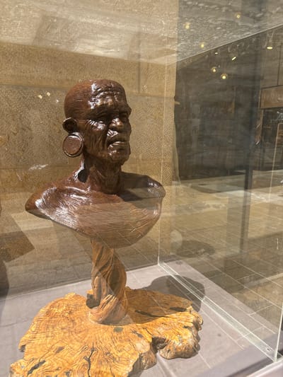 This image features a wooden sculpture of a human head and shoulders, displayed in a glass enclosure. The sculpture, carved from dark, polished wood, depicts a face with deeply etched lines, suggesting age or a weathered expression. A circular object, possibly an earring or a button, is visible on one side of the head. The sculpture is mounted on a rough, textured wooden base, its natural grain and form contrasting with the smooth finish of the head.
The sculpture is positioned within a transparent glass display case, which reflects the surrounding environment. Through the glass, one can discern a tiled wall made of light-colored stone, characteristic of the architecture found in Jerusalem, Israel. The lighting suggests an indoor setting, possibly a museum or gallery, with several small, circular lights visible in the reflection above the sculpture. The overall impression is one of artistic presentation, highlighting the craftsmanship of the wooden carving. There is no visible text in the image.