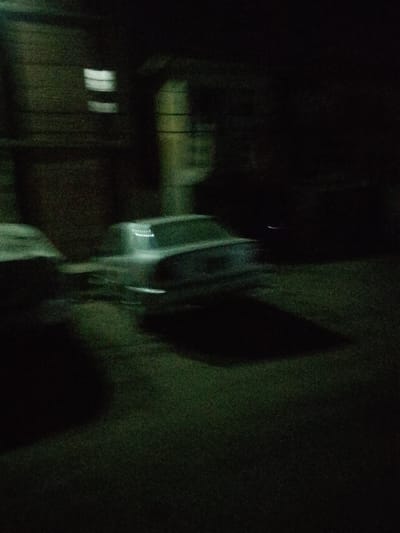 A blurry nighttime shot reveals parked cars and buildings. A white vehicle is positioned in the middle ground with a dark shadow underneath. A building with illuminated windows appears on the left. Another blurry car can be seen on the left side of the frame, almost blending into the dark background. The lighting is dim and unclear, making details difficult to discern. The overall effect is one of darkness and motion blur.