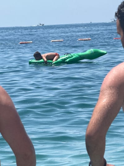 A young boy, wearing red swim trunks, is lying on an inflatable green alligator float in the blue water of the Adriatic Sea. Two people are partially visible in the foreground, their arms and shoulders in the shot, looking out towards the boy. In the background, several buoys mark a swimming area, and a white boat is visible on the horizon. The scene is set in Bečići, Montenegro, a coastal town known for its beautiful beaches and clear waters. The water is calm with gentle waves, and the sky is clear and bright blue.