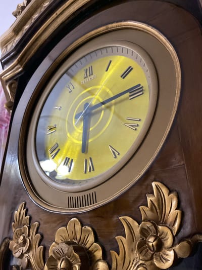 The image shows a close-up, low-angle view of a vintage grandfather clock. The clock face is a bright yellow with Roman numerals and black hands. The word "SEIKO" is visible on the clock face. The clock is housed in a dark wood casing with ornate gold detailing, including floral motifs. The clock is situated in an indoor setting, possibly a home or a shop, with hints of pink fabric in the background. The specific location in Margorejo, Indonesia is not discernible from the image.
