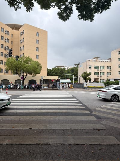 This street scene, likely in Jiangning District, China, shows a crosswalk dividing a street. On both sides, vehicles including cars and scooters are parked. Buildings of light-yellow color with repetitive window patterns line the street. People are visible near what appears to be a bus stop area. The sky is overcast. A traffic light is present. Green trees add foliage to the urban scene. The perspective is slightly tilted.