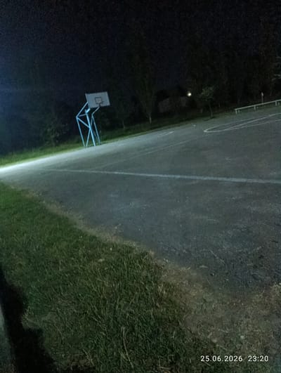 This is a nighttime photograph of an outdoor basketball court. The court surface is asphalt with white lines marking boundaries and a semicircle. A blue metal basketball hoop with a white backboard is visible on the left side of the frame. The area surrounding the court is covered in grass, and trees are present in the background, silhouetted against the dark night sky. The timestamp "25.06.2026 23:20" is displayed in the lower right corner. The image appears to be taken in Slobozia-Dușca, Moldova.