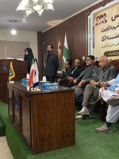 The image captures a gathering, likely a formal event or press conference, taking place indoors. A woman in a hijab is positioned behind a podium, speaking, while a man in a suit stands to her right, appearing attentive. Seated in the foreground are five men, all dressed formally in suits or traditional attire, seemingly listening to the speaker.
Behind them, the Pakistani flag is visible, and next to the podium are smaller flags, one green with a star and crescent. The room is decorated with a banner featuring Arabic script and faces, possibly of revered figures. The environment suggests an official setting, potentially a conference room or a government building. Based on the text within the image, it may be located in Quetta, Pakistan. The scene depicts a formal event involving multiple individuals, with flags and banners suggesting a nationalistic or politically oriented meeting.