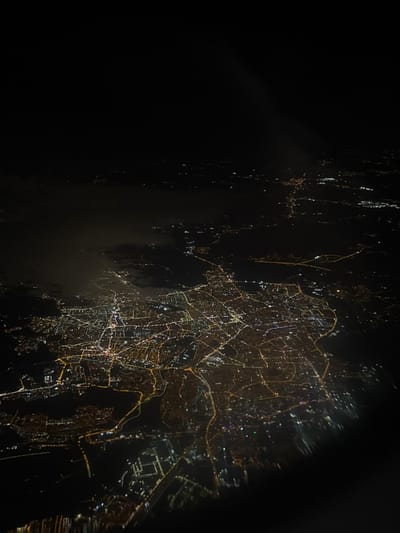 This is an aerial night photograph of the city of Sharjah, United Arab Emirates. The image captures a sprawling urban landscape illuminated by artificial lights, viewed from a high altitude, likely an aircraft. The scene is dominated by a dense network of warm yellow and orange lights, delineating streets, roads, and building clusters that form intricate patterns across the ground. Scattered white and bluish lights are also visible within the more intensely lit urban centers. Dark, irregular masses, possibly low clouds or atmospheric haze, are present over portions of the illuminated areas, particularly in the upper-left and central-left regions, obscuring some ground lights. The areas surrounding the city lights are uniformly dark, suggesting unpopulated land or water bodies. A faint, diagonal light streak is present in the upper-right quadrant, and the lower-right edge exhibits slight blurring consistent with an image taken through an aircraft window. No human figures, specific individual objects, or dynamic actions are discernible from this perspective.