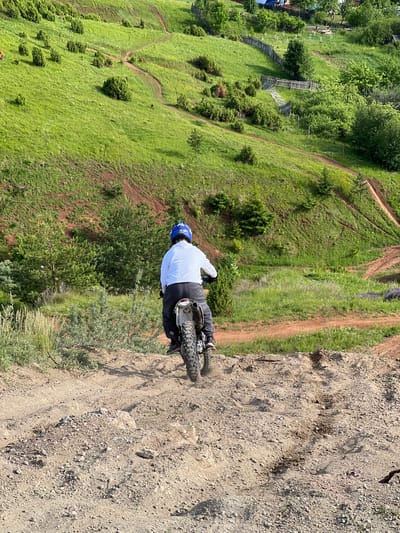 A high-angle shot captures a person riding a dirt bike on a dusty, rocky trail within a hilly, green landscape. The rider is seen from behind, wearing a white long-sleeved top, dark pants, and a bright blue helmet, positioned over the rear wheel of the motorcycle. The dirt bike has knobby tires, characteristic of off-road vehicles. The rear wheel is slightly obscured by dust, indicating motion.
The setting is an open, natural environment with rolling hills covered predominantly in vibrant green grass, interspersed with scattered bushes and small trees. Multiple narrow, winding dirt trails crisscross the hillsides, suggesting a network for off-road activities. The trail the rider is on contrasts sharply with the green, appearing dry, loose, and gravelly.
The bright sunlight suggests it is daytime, likely midday or afternoon, with no visible clouds. The overall impression is one of an active outdoor sport in a rural or semi-wilderness area. The location context indicates this scene is in Ольховка, Russia. There is no visible text in the image.