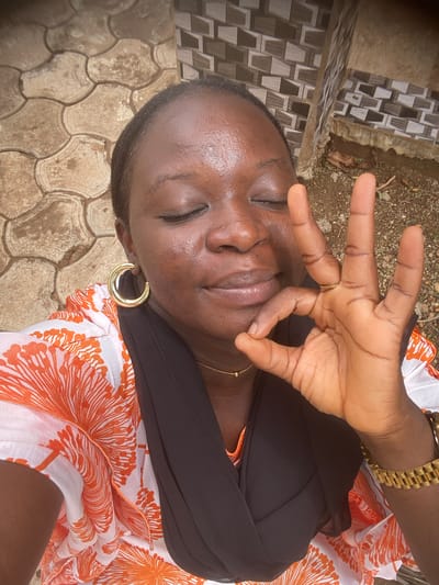 A medium-close, medium-angle photograph shows a dark-skinned woman lying on her back outdoors. She has her eyes closed and is making the "ok" gesture with her right hand over her face. She wears a black scarf or head covering, a white blouse with orange floral designs, a gold wristwatch on her right wrist, a gold ring on her right hand, and gold hoop earrings in her left ear. A gold chain is visible around her neck.
The woman appears to be lying on the ground. The ground is covered in tan paving stones with a geometric pattern. A wall with decorative tiles is visible in the upper-left corner of the image. The background is blurred and shows some ground with sparse vegetation.
The image was taken in Maigizo, Nigeria.