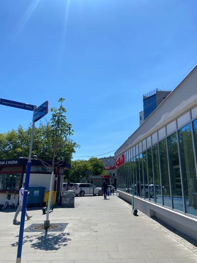 In Warsaw, Poland, under a clear blue sky, a street scene unfolds. A modern building with a glass facade, reflecting the surrounding trees and sky, dominates the right side of the frame. To the left, a blue street signpost stands, with directional signs indicating "Szpital Czerniakowski 450 m" and other distances. Adjacent to the signpost is a small kiosk with a red awning, identified by the text "DZAK Z ROZNA." Several electric scooters are parked nearby, and two people are visible walking on the paved sidewalk. One person, wearing a white t-shirt and grey pants, walks away from the camera, while another, in dark clothing, walks towards the camera carrying a blue bag. In the background, cars are parked, and other buildings and trees complete the urban landscape.