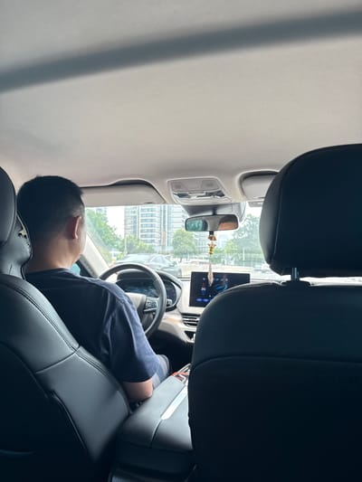 The image shows the interior of a car, taken from the back seat, looking forward. A man is seated in the driver's seat, wearing a dark blue t-shirt. He is looking forward, as if driving or about to drive. The car's dashboard and steering wheel are visible. A digital display screen shows "24.1". Outside the car, through the windshield, a city landscape with tall buildings and trees can be seen. The scene appears to be in Tianhe District, China, given the context of the prompt. There is a decorative item hanging from the rearview mirror. The overall lighting suggests it is daytime.