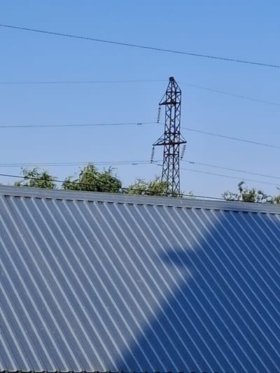 The image shows a power line tower against a clear blue sky. To the right of the power line, a section of a corrugated metal structure, possibly a roof or wall, is visible. The metal surface has a ribbed texture and is reflecting some light, creating a pattern of diagonal lines. There are some green leafy branches growing from the top of the metal structure, reaching towards the power lines. The power line tower is made of dark metal lattice and has insulators from which wires are suspended. The overall scene is an outdoor view with a focus on infrastructure and nature encroaching upon it. The time of day appears to be daytime due to the bright, clear sky. There are no people or explicit activities depicted. The location context is Taraz city administration, Kazakhstan. There is no visible text in the image.