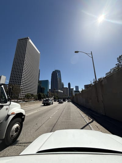 The image depicts a daytime urban scene from a vehicle on a highway, likely within the city of Los Angeles, United States. The view includes a line of cars on a multi-lane highway, skyscrapers, and a clear blue sky with the sun prominently visible. A large white building with a grid-like facade is visible to the left of the road, and other commercial buildings can be seen in the background. Parts of a white truck are visible in the foreground. The highway is concrete, and a concrete barrier separates the road from the adjacent buildings.