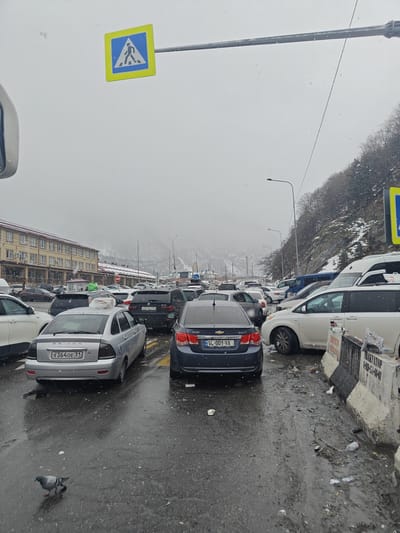 The image depicts a congested street in Verkhnii Lars, Russia, on a snowy day. Numerous cars are seen, with registration plates indicating origins from both Russia and Georgia. A pedestrian crossing sign is visible above the street. The road is wet, indicating recent or ongoing precipitation. Litter and debris are present along the roadside. A single pigeon stands on the street. Buildings are visible in the background, along with a hillside covered with snow-dusted trees.