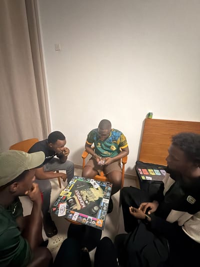 An overhead shot captures a group of at least four young men engaged in a board game session indoors. The central focus is a "MONOPOLY: GAME OF THRONES" edition board game laid out on a low surface, around which the individuals are gathered.
One man, wearing a green and yellow patterned shirt, sits on a wooden chair, looking down at the game or his phone. Another, dressed in a black long-sleeved top and dark pants, is also seated on a chair, with his hands clasped. A third individual, wearing a dark t-shirt and grey pants, leans forward, seemingly focused on the game. Part of a fourth person is visible in the foreground, wearing a green cap and a dark t-shirt. Monopoly money in various denominations is scattered around the game board, suggesting active play.
The setting appears to be a casual living space with plain white walls and a light-colored floor, possibly carpeted or tiled. A wooden cabinet or shelf is visible in the background, with a green drink can resting on it. The lighting is artificial, suggesting the scene takes place in the evening or night. The overall atmosphere is relaxed and focused on the game.
The individuals are dressed casually. One person has "EVIBE..." partially visible on their shirt, and "OUDD..." on a cap. Given the location context provided, this scene is taking place in Jiangning District, China.
