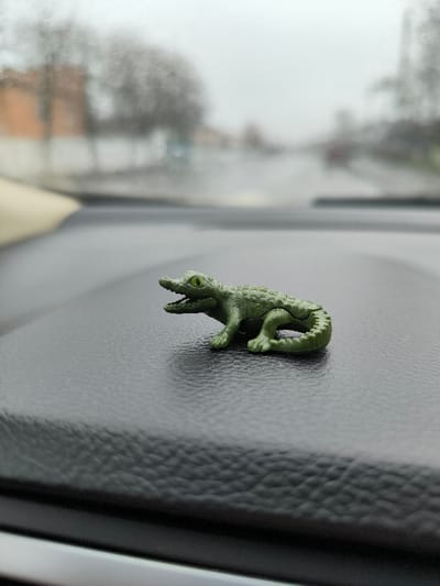 A small, dark green plastic toy depicting an alligator or crocodile with yellow eyes and an open mouth rests on a textured, dark gray vehicle dashboard. The toy faces forward, towards the vehicle's front. Through the windshield, a blurred street scene in Illintsi, Ukraine, is visible under an overcast, light gray sky. Bare trees and indistinct buildings line the sides of the road, which recedes into the distance. No individuals or active subjects are present in the frame.