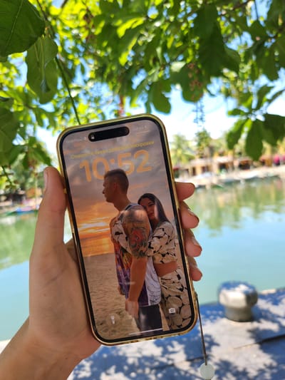 In Hội An Ward, Vietnam, a hand holds up a smartphone displaying a lock screen with a picture of a couple embracing on a beach during sunset. The phone's screen shows the time as 10:52 and the text "Смахните вверх для разблокировки" which translates to "Swipe up to unlock" in Russian. The background of the image is a blurred view of a body of water, likely a river or canal, with buildings and greenery visible on the opposite bank. The overall scene suggests a sunny day, possibly during a vacation or leisure activity.