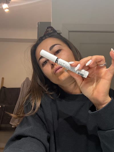 A woman with long brown hair and a black sweatshirt holds a white tube with black writing up to the camera. She has a ring on her left hand, and her nails are painted white. A hair clip is visible in her hair. The background shows a door, wall, and what appears to be a towel hanging on a rack. The image lacks details that would allow for identification of a location in Cannes, France.