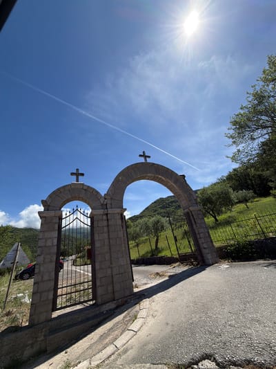 The image displays a rustic stone gateway with two arched openings, situated within a scenic, hilly landscape. Each arch of the gateway is crowned with a simple cross, suggesting religious or historical significance, possibly marking the entrance to a sacred site or private property. The structure is built from rough-hewn stone blocks, and dark iron gates are fitted within the arched openings.
To the right of the gateway, a paved asphalt road runs parallel, curving gently out of frame. Beyond the road and extending from the gateway, a metal fence encloses a lush, green area abundant with trees and foliage, typical of a natural, rural environment. Through the openwork of the gates and in the background, verdant, tree-covered mountains or hills rise under a clear blue sky.
The sky is bright and expansive, with the sun radiating intensely from the upper left, creating a prominent sunburst effect. A long, thin contrail stretches horizontally across the sky, accompanied by a few faint, wispy clouds. A small part of a dark-colored car is visible through the right gate, near the bottom. The scene is bathed in bright daylight, indicating either morning or afternoon.
This tranquil setting is located in Kubasi, Montenegro, characterized by its natural beauty and traditional architecture. There are no people visible, nor any specific activity taking place. No discernible text is present in the image.