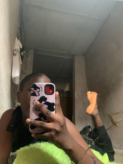 The image is a mirror selfie captured indoors in Agege, Nigeria. A person with dark skin, wearing a dark top, is depicted reclining, partially obscuring their face with a vertically held smartphone. The phone's light pink case features black bunny designs with bows and text including "LUCKY RABBIT" and "THANK YOU". The phone displays a red camera lens. A small silver earring is visible in the individual's left ear. In the foreground, a bright green, textured textile object supports the individual's right arm. To the right, a bare left foot, likely belonging to the same individual, is visible with the sole facing the viewer. The room's background shows unpainted, light grey/brown textured walls with visible seams, and a light-colored panelled ceiling. To the left, a white shirt and a dark garment hang on a wall hook. A dark, rectangular doorway or opening is visible centrally in the background.