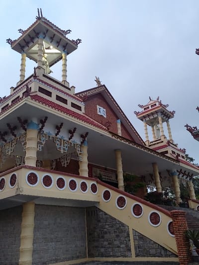 The image displays the exterior of the Domaine de Marie Church in Thành phố Đà Lạt, Vietnam. The multi-tiered structure exhibits a blend of traditional Vietnamese and French colonial architectural styles. The building features prominent red-tiled roofs with ornate, upward-curving eaves and decorative finials. A three-tiered tower on the left side, topped by a supported canopy, houses a white statue of Jesus Christ with outstretched arms. The main building's red brick gable section prominently displays the white Greek letters "Α Ω" (Alpha and Omega). A wide, cream-colored veranda is visible, featuring a balustrade with white circular decorative cutouts and supported by light-colored columns. An ascending staircase with a matching cream-colored balustrade and circular cutouts leads to this veranda. The base of the building and the lower section of the staircase are constructed from grey, horizontally-laid stone blocks. The sky is overcast. No individuals are present in the scene.