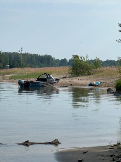 A serene waterscape captures a light blue sky above a tranquil body of water. The water reflects the sky and the surrounding environment, which includes a sandy beach lined with tall, dry grass and scattered green bushes and trees in the background.
Dominating the foreground of the water is a dark-colored motorboat, moored near the shore and partially submerged. The boat has a windshield and appears to be equipped with fishing rods. Further down the beach, a blue inflatable tube lies on the sand, next to some other objects, including a dark bag and what appears to be a piece of blue plastic sheeting.
The beach itself is a light brown sand with some scattered debris, including driftwood and small rocks. The overall impression is one of a quiet, natural setting on a clear day. The image does not contain any visible people. The location is identified as Beregovoe, Russia.