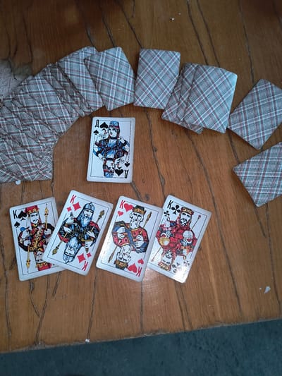 The image shows a spread of playing cards on a wooden surface. The cards are laid out in a fanned pattern, with some face up and others face down. The face-up cards include three Kings and one Ace. The Kings are all depicted with beards and regal attire, each holding a scepter and a orb. The Ace is of spades and features a woman in blue and red attire. The face-down cards have a red and blue plaid pattern on their backs. The wooden surface appears to be a table with visible grain and some wear. There are no people visible in the image, and no specific landmarks or clues are present to indicate a location in Rivne, Ukraine.