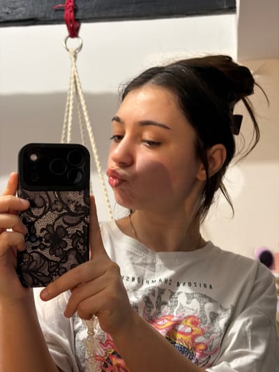 An indoor mirror selfie captures a young woman from the chest up, reflected in a mirror or reflective surface. The image is oriented with the woman's head to the bottom right and her body extending towards the top left. She has dark hair pulled back, with some loose strands around her face, and appears to be making a playful "fish face" or puckering her lips. She is wearing a white short-sleeved t-shirt with a graphic design and mirrored text that reads "ARIZONA CHAMPIONS" or "ARIZONA CHAMPIONSHIP." A thin gold-colored necklace is visible around her neck.

She holds a dark-colored smartphone, likely an iPhone, with a distinct black lace or floral patterned case. Her hands are visible holding the phone, which obscures part of her lower body in the reflection.

The background behind her shows a plain light-colored wall. To her left (viewer's left), a dark vertical structure, possibly a wooden beam or frame, is visible. Red string or fabric is tied around this structure, from which a silver ring hangs, attached to a light-colored, braided macrame-style string that extends out of view. The setting appears to be a casual indoor environment. The time of day is not explicitly indicated, but the lighting suggests a well-lit interior. Her expression conveys a sense of lightheartedness or fun.