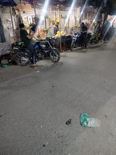 The scene is a nighttime street in Dhaka, Bangladesh, at latitude 23.784 and longitude 90.377. A man is sitting on a motorcycle, facing the camera, holding a helmet in his right hand. He is wearing a dark blue shirt and black pants. The motorcycle is parked on the side of the street. A second motorcycle is parked across the street, and several people are walking away from the camera. The street is lit by streetlights and appears to be in front of a row of street vendors. There are two plastic bags lying on the street.