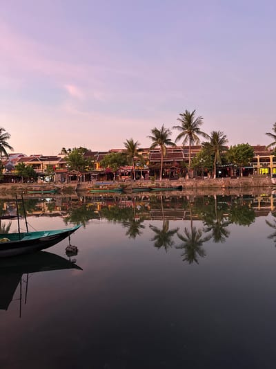 The image depicts a tranquil riverside scene in Hoi An, Vietnam, likely during twilight. The sky displays a gradient of soft pink and purple hues. Along the riverbank, a row of traditional Vietnamese buildings with tiled roofs and colorful facades stands, interspersed with lush green trees and palm trees. Several boats are moored or gently floating on the water. In the foreground, a green and black boat with a pole is visible, its reflection perfectly mirrored in the still water, along with the reflections of the palm trees and buildings. The overall atmosphere is serene and picturesque, showcasing the charm of Hoi An.